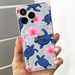 Pode incluir: Capa de telemóvel transparente com um padrão de tartarugas marinhas azuis e flores cor-de-rosa. A capa tem um porta-cartões transparente. O design inclui pequenos pontos azuis num fundo azul claro.