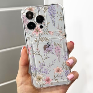 Pode incluir: Uma capa de telemóvel transparente com um padrão floral delicado de lírios cor-de-rosa e flores de glicínia roxas. O design inclui caules castanhos finos e folhas verdes impressas na superfície transparente.