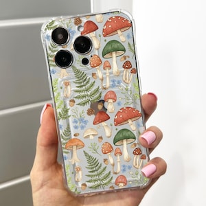 May include: Clear phone case with a whimsical mushroom pattern. The design features various mushrooms in shades of red, green, and brown, along with ferns, blue flowers, and acorns. The case is held in a hand with pink nail polish.