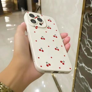 May include: A white phone case with a cherry pattern. The case has a raised camera lens cover with three holes for the camera lenses.