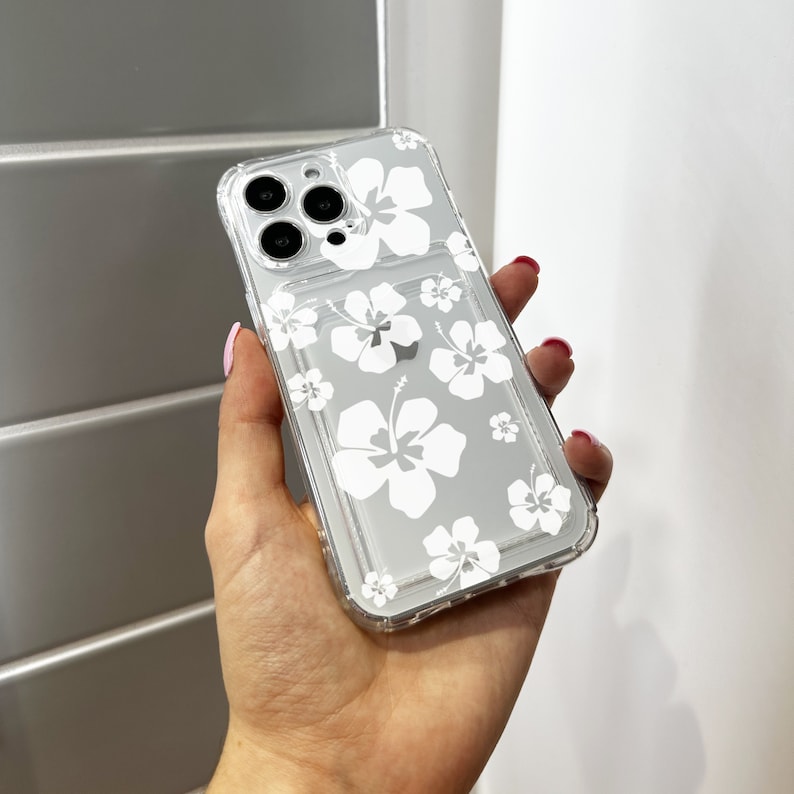 May include: A clear phone case with a white hibiscus flower pattern. The case is designed for a smartphone with a triple camera lens.