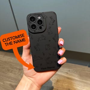 May include: Black phone case with a repeating pattern of rabbit illustrations. The case has a camera cutout and the text "Your Name" at the bottom. An orange sign with the text "CUSTOMISE THE NAME" is in the image.