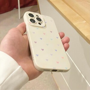 May include: A cream phone case decorated with small, pastel-coloured hearts. The case has a camera cut-out and is held in a hand. The hearts are in shades of pink, purple, and blue.