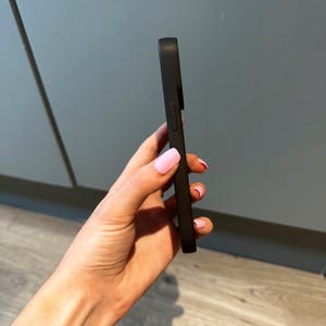 May include: A black phone case, held at an angle, showing the side profile. The case has a smooth, matte finish. The case is designed to protect a smartphone. The case is solid black.