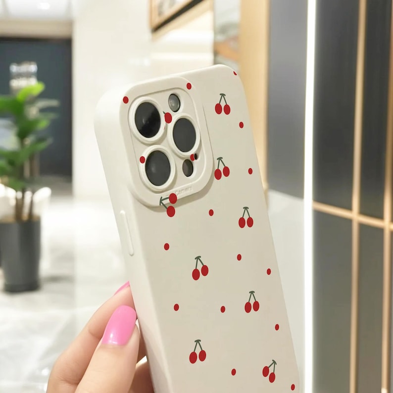 May include: White phone case with a pattern of red cherries. The case has a raised camera lens cover.