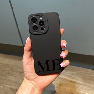 May include: Black phone case with a camera lens cover and the initials "MB" printed on the back.