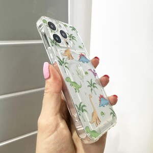 Op de afbeelding: Transparante telefoonhoes met een herhaald patroon van cartoon dinosaurussen en palmbomen. De hoes is gemaakt van transparant plastic, waardoor de kleur van de telefoon zichtbaar is. Het ontwerp bevat verschillende dinosaurussoorten in groen, blauw en oranje.