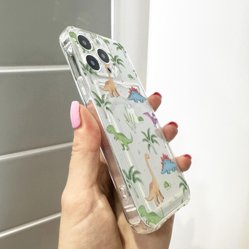 Op de afbeelding: Transparante telefoonhoes met een herhaald patroon van cartoon dinosaurussen en palmbomen. De hoes is gemaakt van transparant plastic, waardoor de kleur van de telefoon zichtbaar is. Het ontwerp bevat verschillende dinosaurussoorten in groen, blauw en oranje.
