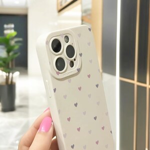May include: White phone case with a pattern of small purple and pink hearts. The case has a raised camera lens cover with three black camera lenses.