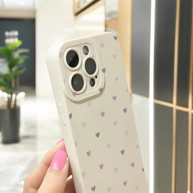 May include: White phone case with a pattern of small purple and pink hearts. The case has a raised camera lens cover with three black camera lenses.