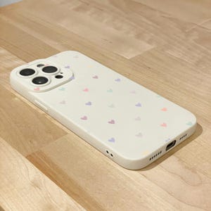 May include: A cream-coloured phone case decorated with small, pastel-coloured hearts. The case features a raised camera module and cutouts for ports and speakers. The hearts are in shades of pink, purple, blue, and orange.