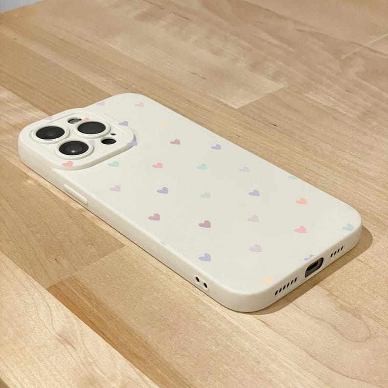 May include: A cream-coloured phone case decorated with small, pastel-coloured hearts. The case features a raised camera module and cutouts for ports and speakers. The hearts are in shades of pink, purple, blue, and orange.