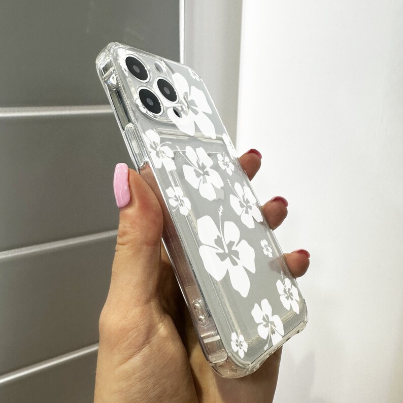 May include: A clear phone case with a white hibiscus flower design. The case is made of a flexible material and has a raised lip around the camera lens to protect it from scratches.