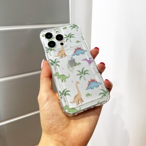 Op de afbeelding: Transparante telefoonhoes met een kleurrijk dinosauruspatroon. De hoes heeft verschillende dinosaurussen, palmbomen en andere elementen. De hoes heeft een kaarthouder op de achterkant.