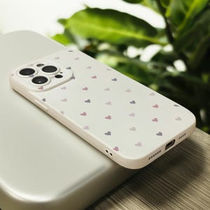 May include: White phone case with a pattern of small pink and purple hearts. The case has a camera lens cutout with three black circles.