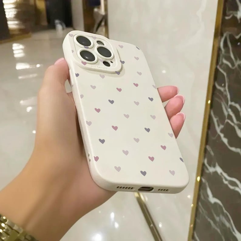 May include: A white phone case with a pattern of small pink and purple hearts. The case has a raised camera lens cover.