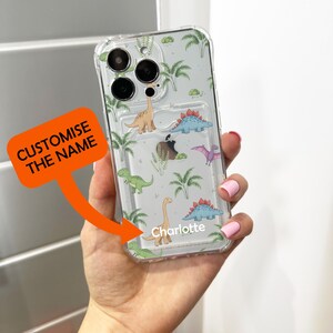 Op de afbeelding: Transparante telefoonhoes met een dinosauruspatroon. De hoes heeft verschillende kleurrijke dinosaurussen en palmbomen op een lichtgrijze achtergrond. De naam "Charlotte" staat op de hoes. De tekst "CUSTOMISE THE NAME" staat op een oranje label.