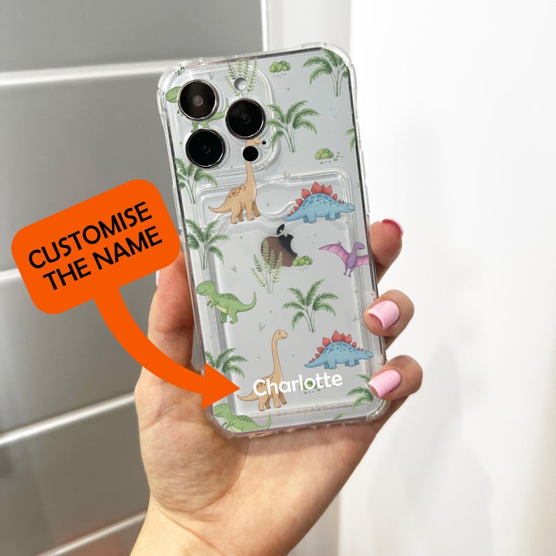 Op de afbeelding: Transparante telefoonhoes met een dinosauruspatroon. De hoes heeft verschillende kleurrijke dinosaurussen en palmbomen op een lichtgrijze achtergrond. De naam "Charlotte" staat op de hoes. De tekst "CUSTOMISE THE NAME" staat op een oranje label.