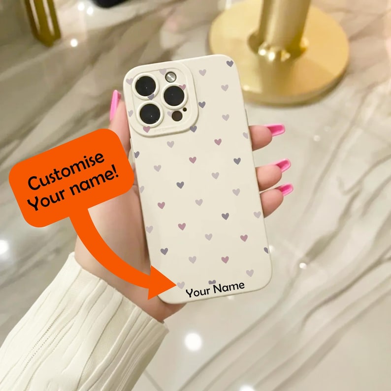 May include: A white phone case with a pattern of small pink and purple hearts. The case has a cutout for the camera lens and a space at the bottom for a personalized name. The text "Your Name" is printed on the case.