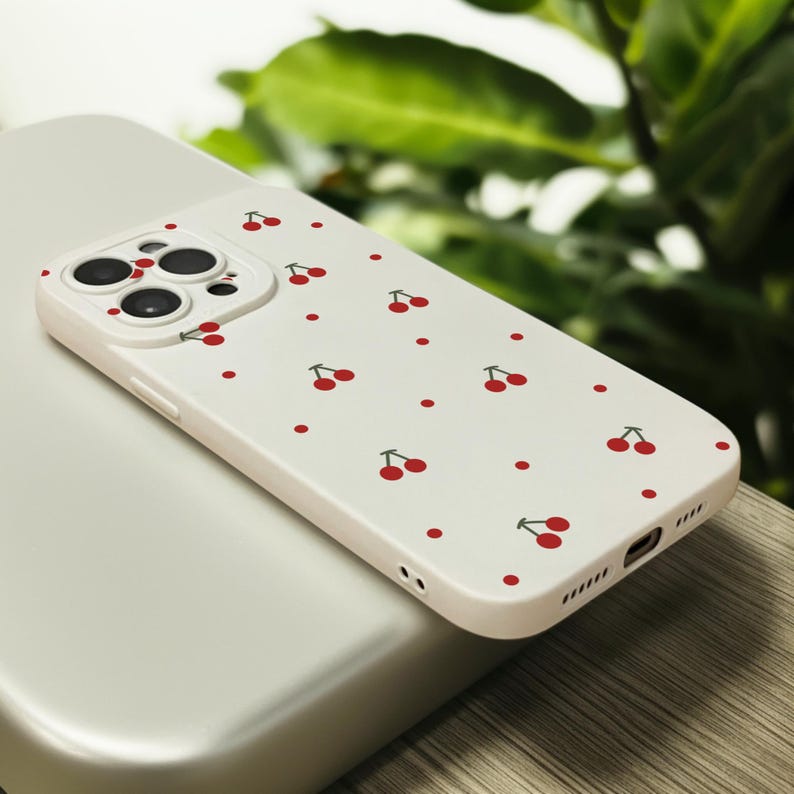 May include: A white phone case with a pattern of red cherries and white dots. The case has a raised camera bump with three lenses.