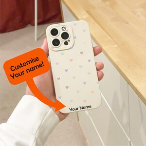 May include: A cream phone case decorated with small pastel hearts. The case has a camera cutout and the text "Your Name" at the bottom. An orange speech bubble says "Customise Your name!"