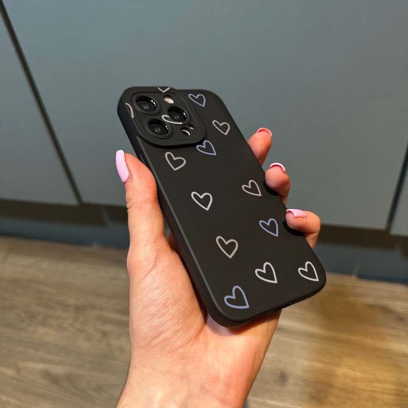 Puede incluir: Carcasa de tel&eacute;fono negra con un patr&oacute;n de corazones blancos. La carcasa tiene una cubierta de lente de c&aacute;mara con tres agujeros.