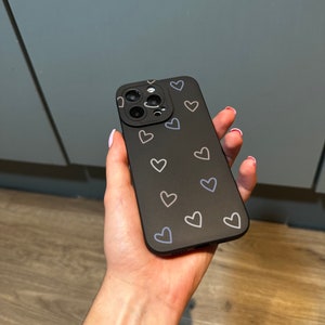 Puede incluir: Carcasa de tel&eacute;fono negra con un patr&oacute;n de corazones blancos. La carcasa tiene una protuberancia de c&aacute;mara elevada con tres lentes.