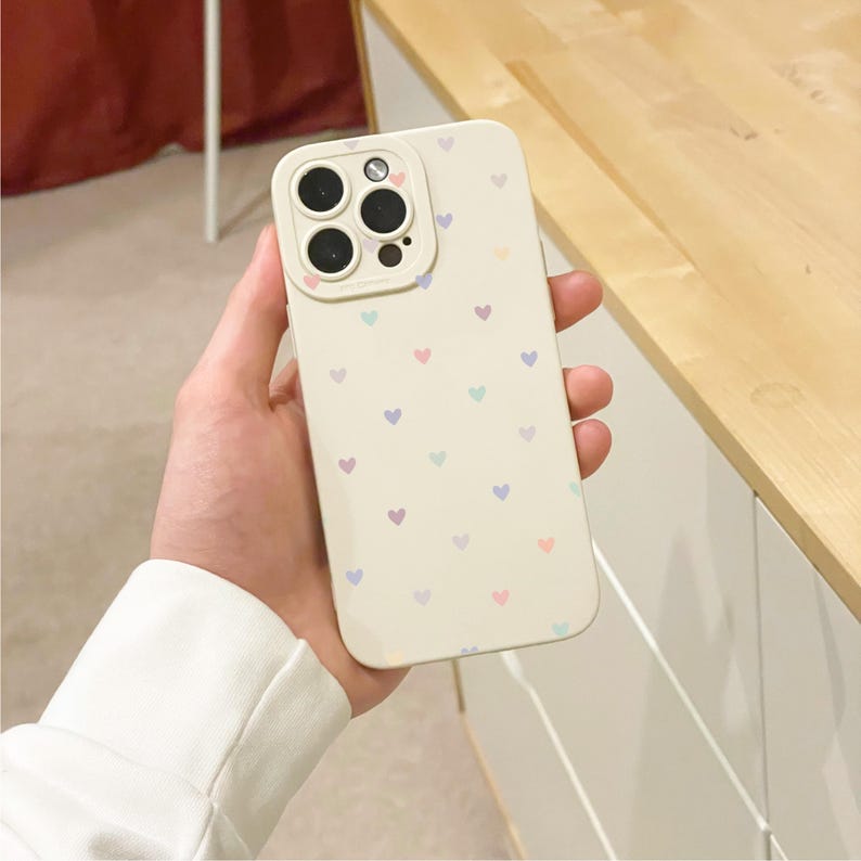 May include: A cream phone case decorated with a pattern of small, pastel-coloured hearts. The case has a protective camera lens surround. The case is held in a hand, showing the design and fit.