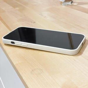 May include: A white phone case with a black screen sits on a light-coloured wooden surface. The case has rounded edges and a modern design. The phone's screen is dark, suggesting it is either off or displaying a black image.