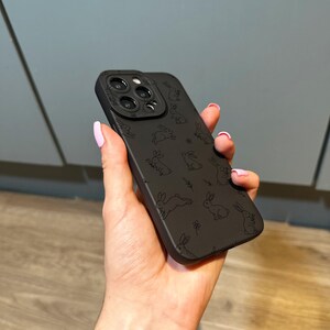 May include: A black phone case featuring a repeating pattern of sketched rabbits and small floral accents. The case has a camera lens protector and is held in a hand with pink nail polish.
