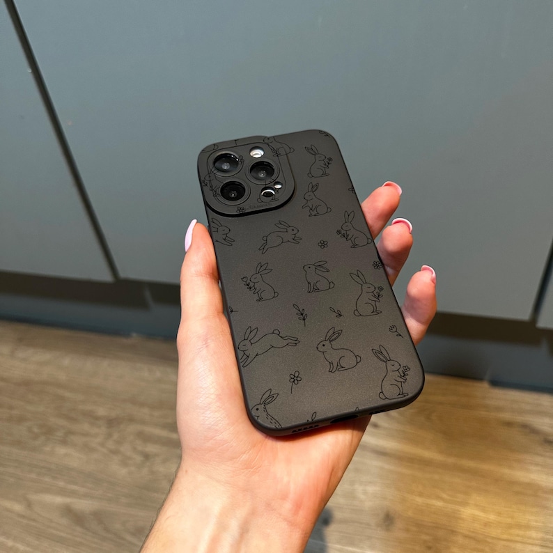 May include: Black phone case with a repeating pattern of sketched rabbits and small floral accents. The case has cutouts for the phone's camera and buttons. The overall design is minimalist and features a matte finish.