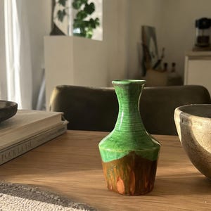 Può includere: Un piccolo vaso decorativo in ceramica, verde nella parte superiore e marrone in quella inferiore. Il vaso ha un design strutturato e a coste, con un collo svasato. È appoggiato su un tavolo di legno, accanto a una ciotola e libri.