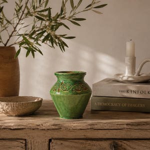 Può includere: Una natura morta con un vaso di ceramica verde decorato, una piccola ciotola, un vaso di terracotta con rami d'ulivo, un portacandele e due libri. Gli oggetti sono disposti su una superficie di legno.
