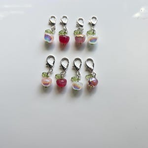 Può includere: Otto ciondoli a forma di mela con chiusure color argento. Le mele sono un mix di colori rosa, rosso e iridescenti, ognuna con una foglia e un gambo verdi. I ciondoli sono disposti in due file da quattro su sfondo bianco.