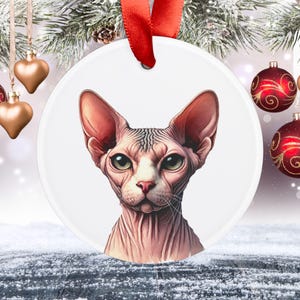 Puede incluir: Adorno navideño redondo y blanco con una ilustración detallada de la cara de un gato Sphynx. El gato tiene piel rosada, orejas grandes y ojos verdes. Se adjunta una cinta roja para colgarlo. Decoración festiva.