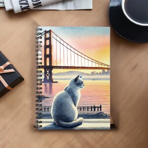 Peut inclure: Un chat blanc est assis sur une jetée surplombant le Golden Gate Bridge à San Francisco. Le pont est en arrière-plan et l'eau au premier plan. Le ciel est d'un orange et d'un rose vifs.