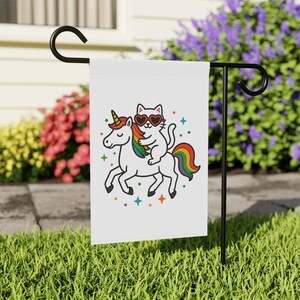 May include: White garden flag featuring a cartoon cat wearing heart-shaped sunglasses riding a unicorn with a rainbow mane and tail. Colorful stars and accents surround the whimsical design. The flag is displayed on a black metal stand in a grassy yard.