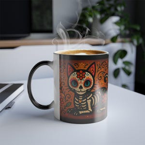 May include: A ceramic coffee mug with a Day of the Dead cat design. The cat has a skeleton body, floral accents, and blue eyes. The mug is filled with a hot drink, and steam rises from the top.