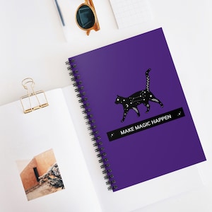 May include: A spiral-bound notebook with a vibrant purple cover. A black cat silhouette with celestial designs is featured, along with the text "MAKE MAGIC HAPPEN" on a black banner. The notebook is open to reveal blank pages.