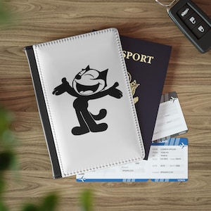 Könnte beinhalten: Ein weißer Reisepasshalter mit einem schwarz-weißen Cartoon-Katzen-Design, neben einem dunkelblauen Reisepass, Flugtickets und einem Autoschlüsselanhänger. Der Reisepasshalter hat einen schwarzen Rücken.