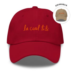 May include: A red baseball cap with the phrase "la cool titi" embroidered in orange script across the front. A smaller image shows a similar cap in a tan color. The cap has a curved brim and a fabric strap.