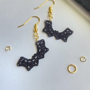 May include: A pair of black lace earrings with gold hooks. The earrings are shaped like a crescent moon and have a delicate, intricate design.