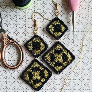 May include: A pair of black and gold crocheted earrings with gold hooks. The earrings are made of two square crocheted pieces, one smaller than the other, with a gold chain connecting them.