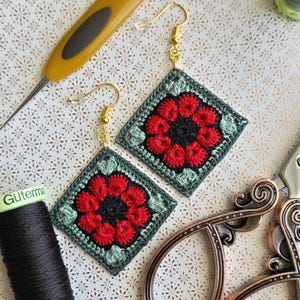 May include: Handmade crochet earrings with a floral design. Each earring features a red and black flower center surrounded by green and gray squares, suspended from gold-colored hooks. The earrings are displayed with crochet tools and black thread.