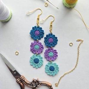May include: Handmade crochet flower earrings in shades of blue, purple, and teal. The earrings feature three layered flower designs with gold-colored ear wires. Crafting supplies, including scissors, thread, and a crochet hook, are arranged around the earrings.