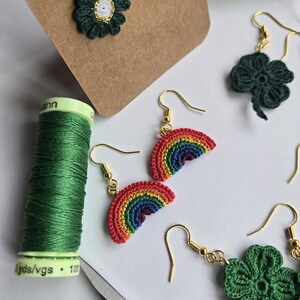 May include: A collection of handmade crochet earrings in various designs. The earrings include green flower shapes with white centers, rainbow arcs, and four-leaf clovers. Gold-colored earring hooks are attached. A spool of green thread and scissors are also visible.