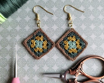 May include: A pair of crocheted earrings with a geometric pattern in shades of green, brown, and yellow. The earrings are hanging from gold hooks.