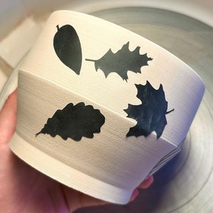 May include: A white ceramic bowl with black leaf silhouettes. The bowl has a textured surface and is slightly tilted. The leaves are in various shapes and sizes, including oak leaves and maple leaves.