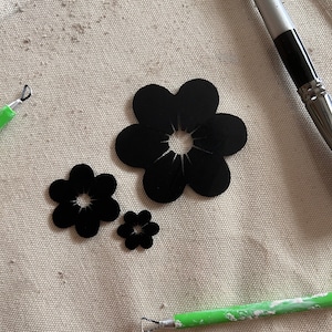 May include: Three black paper flower shapes on a beige fabric surface. The largest flower has a center hole. Two smaller flowers are to the left of the large flower. Two green tools are on the surface.