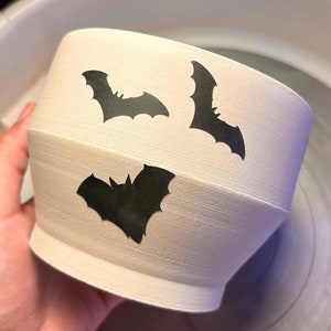 May include: White ceramic planter with three black bat silhouettes. The planter is round and has a wide opening.